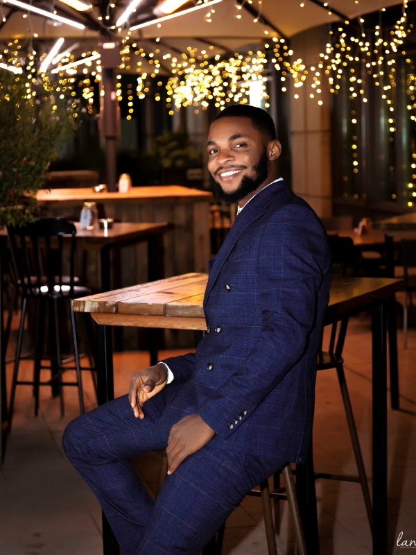 A man in a blue textured suit sitting at a wooden table in an outdoor restaurant decorated with fairy lights. — portrait photography London by Lamss Media